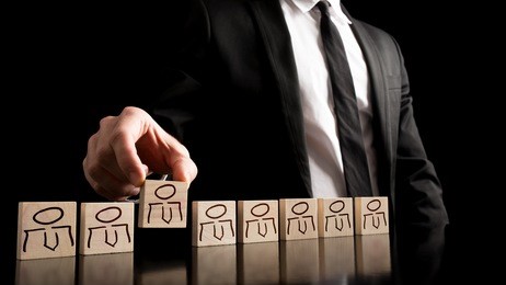 businessman in black and white business suit arranging wooden pieces with people drawings on the table with pure black background. simple human resource concept.