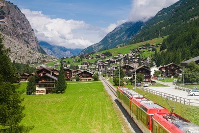red (glacier express) train heading through high mountain valley in switzerland alps on route from st. moritz to zermatt.