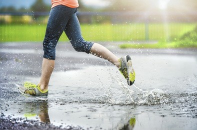 young woman running on asphalt sports field in rainy weather. details of legs and sports shoes splashing in puddles.