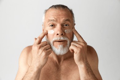 portrait of handsome, mature, gray-haired, bearded man applying face moisturizing cream against grey background. concept of male beauty, face and skin care, daily procedures, age, cosmetics