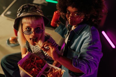 two cool girl friends are outside, sitting on the hood of their car, feeding each other with french fries.