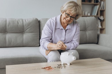 savings, money, annuity insurance, retirement and people concept - smiling senior woman putting coins into piggy bank at home