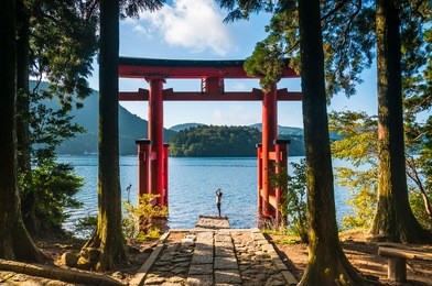 a young woman takes photos with her phone of a torii gate in hakone, japan.
