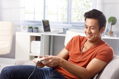 young asian man sitting in armchair at home, playing video game, smiing.