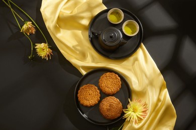 top view of mooncakes displayed on round dishes with tea set, yellow flower and yellow silk cloth on black background. scene for advertising. traditional chinese mid-autumn festival