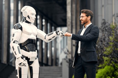 a male businessman in a business suit standing with a robot against the background of a building, shaking hands with each other, fist into fist. the future with artificial intelligence.
