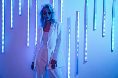 young beautiful hipster woman in street style fashion concept. hot model wearing white suit and sunglasses. female posing in blue neon light in studio interior