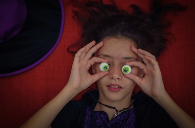 a teenage girl in a witch costume closes her eyes with human eye-shaped candy, lying on an orange-red couch with a halloween hat, close-up top view.