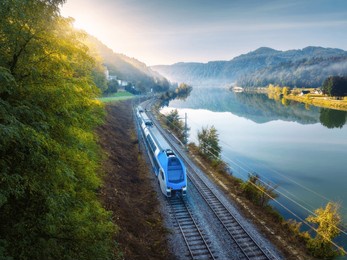 aerial view of blue modern high speed train moving near river in alpine mountains in fog at sunrise in autumn. top view of train, railroad, lake, reflection, trees in fall. railway station in slovenia