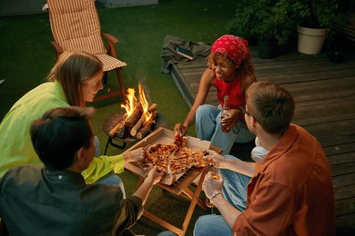 happy friends enjoying pizza and beer drink nearby burning campfire