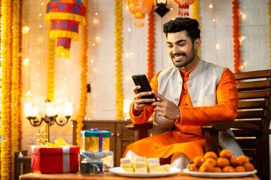 happy young indian man using mobile phone during diwali festival celebration at home in front of sweets and gift boxes on decorated home - concept of festival offers, diwali greetings and social media