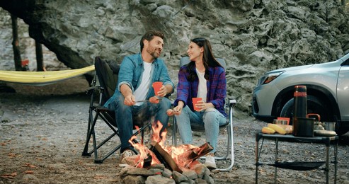 attaractive young woman and bearded man sitting in forest near rock, roasting sausages by bonfire in summer evening, talking, hugging and smiling drinking. time to travel concept. family travel dream