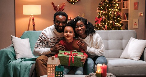 portrait of cheerful african american mom and dad talking and smiling hugging their cute little child son with xmas gift in decorated room with glowing tree at home, smiling to camera. christmas eve