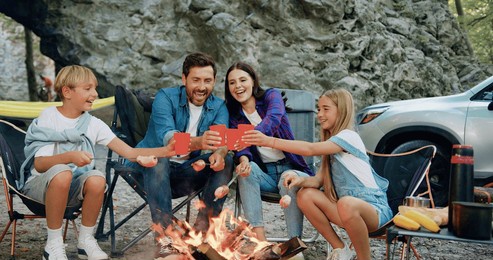 attaractive family mother and father sitting in forest near rock, roasting sausages by bonfire in summer evening, talking, hugging and smiling drinking. time to travel concept. family travel dream