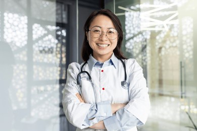 portrait of a young beautiful and successful asian woman inside a clinic office, a doctor smiling and looking at the camera with crossed arms and a white medical coat with a stethoscope.