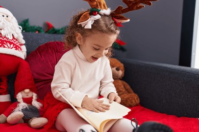 adorable blonde girl reading book sitting on sofa by christmas decor at home