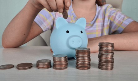 little girl puts coin into blue piggy bank sitting at table at home. child saving money for future, insurance and investment concept