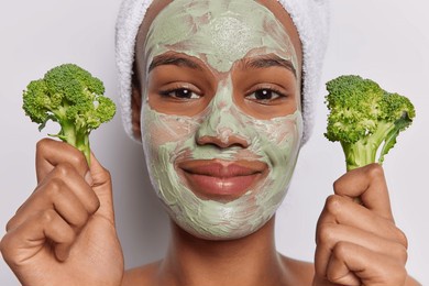 close up shot of pleased young woman applies natural beauty mask holds broccoli undergoes skin care procedures poses with towel wrapped on head isolated over white background. spa procedures