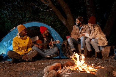 multicultural group of happy friends camping having fun and enjoying their trip in the mountain. black man playing the guitar for his colleagues at bonfire night in the forest.