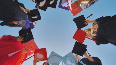 graduates tossing multicolored hats against a blue sky.