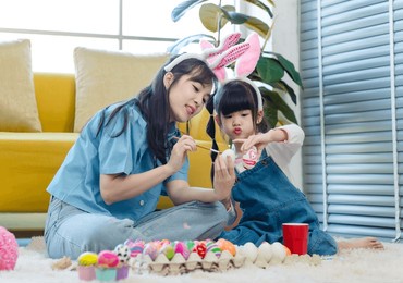 asian cute little children girl wearing funny bunny ears headbands and young pretty mother smile decorating painting eggs while sitting on floor in living room, family preparing for easter holiday.