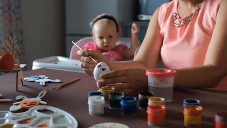 young mother painting easter egg while her cute baby playing in the high chair, selective focus