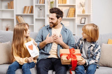 celebrating father's day, a small son and daughter surprise their father with gifts as he sits comfortably on the living room couch, showcasing a scene of pure family joy and contentment