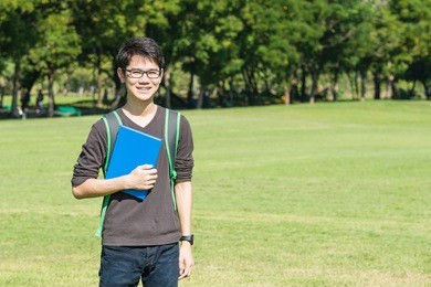 asian student holding books and smiling while standing in park at college