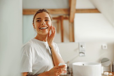 beautiful young woman taking care of skin by applying moisturizer cream in bathroom