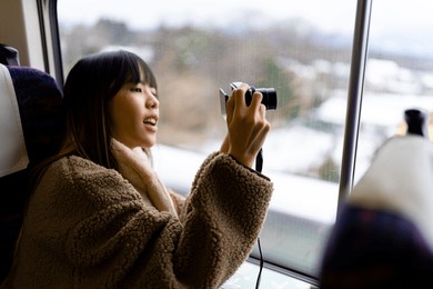 young asian woman using digital camera taking picture of beautiful view out of the window during travel on train at sunset. attractive girl travel japan on railroad transportation on winter vacation.