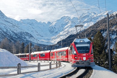 a bernina express travels thru montebello curve on a sunny winter day, with morteratsch glacier lying below piz bernina and snowy mountains in background in pontresina, graubünden, switzerland, europe