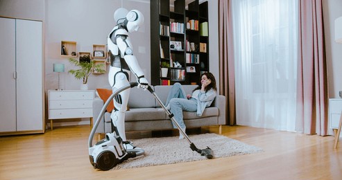 futuristic cyborg using vacuum cleaner while woman in casual wear lying on comfy sofa and talking on smartphone at home. concept of robot and human cooperation.