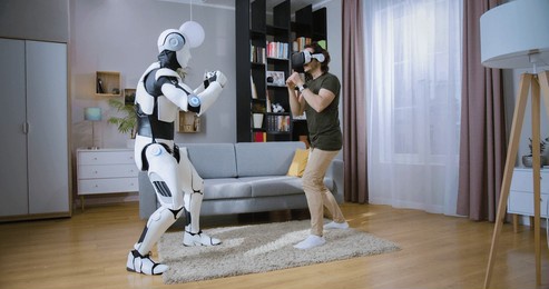 young caucasian man in vr headset boxing with cyborg in cozy living room. handsome male in casual wear having fun with bionic automated robot at home during weekends.