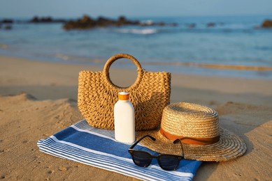 beach scene with straw sun hat, sunscreen bottle, sunglasses, on towel beside ocean. essential sun protection items for skin care during a summer day, placed on sandy shore near sea water.