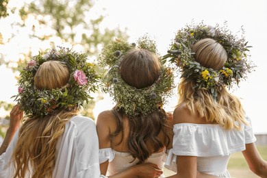 young women wearing wreaths made of beautiful flowers outdoors on sunny day, back view