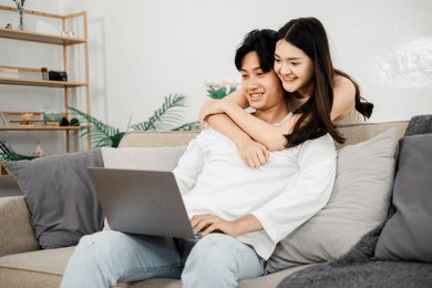 joyful asian couple is engaged with a laptop on their sofa, sharing a happy moment in a comfortable living room.