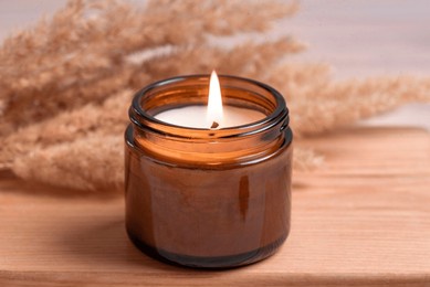 cozy burning candle in amber glass jar, winter hygge home interior decor on wooden tray with dry reeds or pampas grass close up. scented wax eco candle.