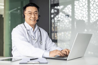 professional asian male doctor with a stethoscope engaging in an online consultation, using a laptop in a well-lit office.
