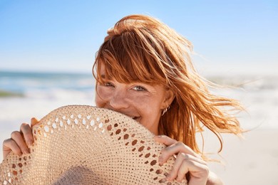 portrait of beautiful woman hiding behind a straw hat at beach and looking at camera with beautiful green eyes. closeup face of smiling girl with freckles and red hair relaxing at seaside.