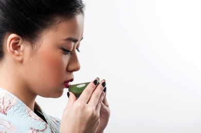 enjoying tea ceremony. side view portrait of young beautiful japanese woman in kimono drinking tea with copy space while standing against white background