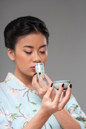 asian beauty drinking tea. low angle portrait with selective focus of young beautiful japanese woman in traditional kimono drinking tea during tea ceremony while standing against grey background