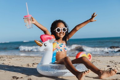 cheerful young girl with sunglasses and unicorn float relaxing at the beach, holding a drink, and having fun during a summer vacation.
