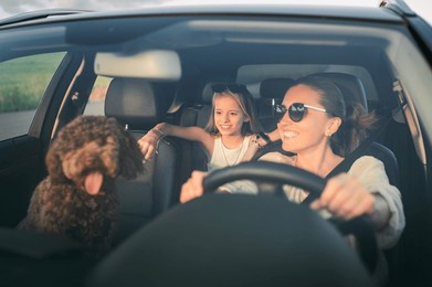 smiling woman driving car with  brown maltipoo dog and her daughter in backseat, enjoying sunny day. image captures joyful family moment during road trip, highlighting happiness and togetherness