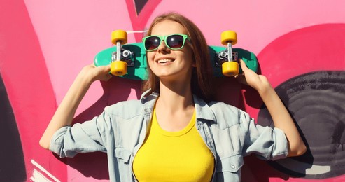stylish happy young girl with skateboard in glasses, modern teenager smiling girl looking away posing against the background of pink graffiti wall