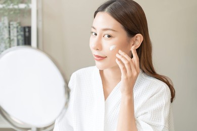 facial beauty skin care, smile of pretty asian young woman in bathrobe looking at mirror, hand applying moisturizer lotion on her face, putting cream treatment before makeup cosmetic routine at home.