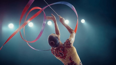 asian female gymnast elegantly twirls vibrant ribbon under bright stadium lights, her precision, grace, and artistry in captivating rhythmic gymnastics performance.