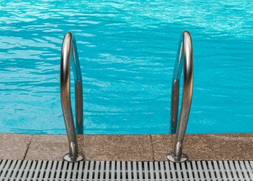 a close-up view of the stainless steel handrails of a swimming pool. the handrails are secured to the deck, which is made of large, brown tiles. the pool is filled with sparkling blue water.
