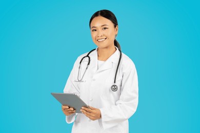 asian woman doctor wearing a white lab coat and stethoscope is smiling while holding a tablet in front of a bright blue background.