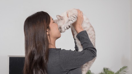 a woman standing in a living room, holding her tortoiseshell cat close to her chest. the setting is calm and domestic