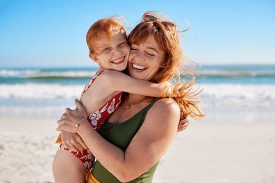 happy young woman embracing her daughter at beach with copy space while laughing together. cute little girl with freckles playing with mom outdoor at seaside. close up of carefree mom hugging child.